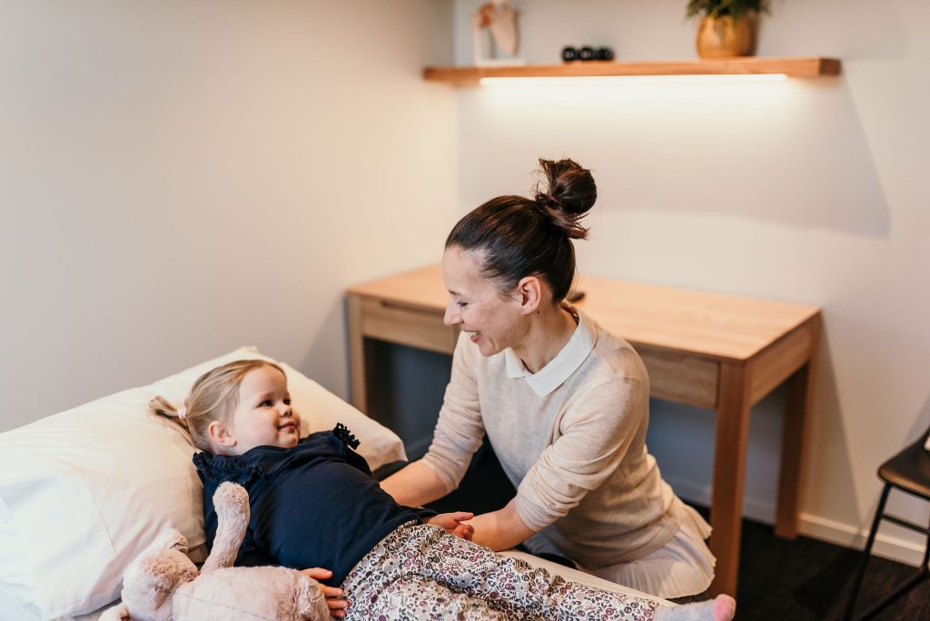 Osteopath smiling while gently interacting with a young child lying on a treatment table, with a plush toy by her side.